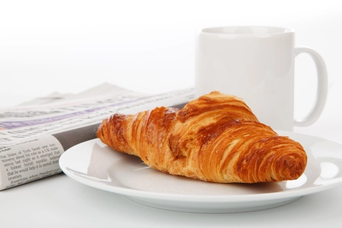 Foto di un croissant accanto a una tazza bianca e a un quotidiano.