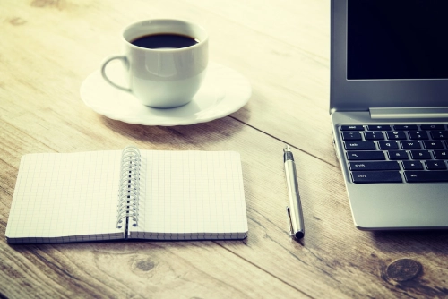 Foto di un taccuino, una penna, una tazza di caffè e un pc sopra una scrivania di legno.