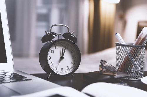 Foto di un orologio a sveglia sulla scrivania accanto a un pc e a un portapenne.