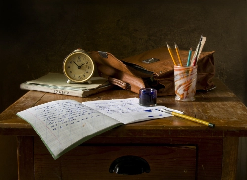 Foto di un quaderno, una matita, un portapenne, una sveglia, un libro e una valigetta di pelle marrone sopra una scrivania di legno antica.