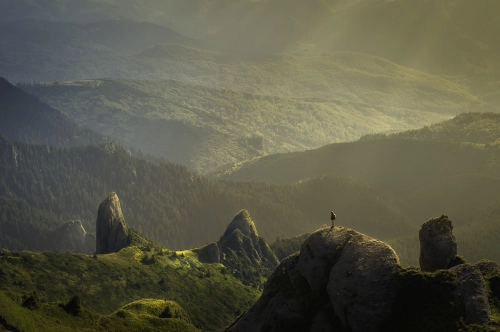 Veduta aerea di un paesaggio montano e boscoso, con una persona ritratta in lontananza sulla cima di una rupe.