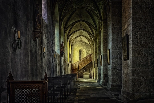 Dipinto di una cattedrale medievale gotica, in penombra e con una luce sotto le volte a sesto acuto.